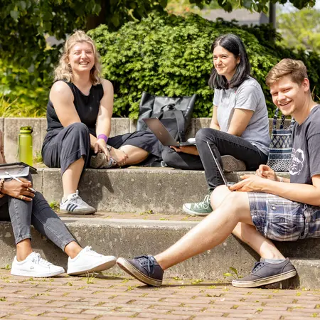 Vier junge Erwachsene draußen auf einer Treppe des Hochschulgeländes mit Laptops 