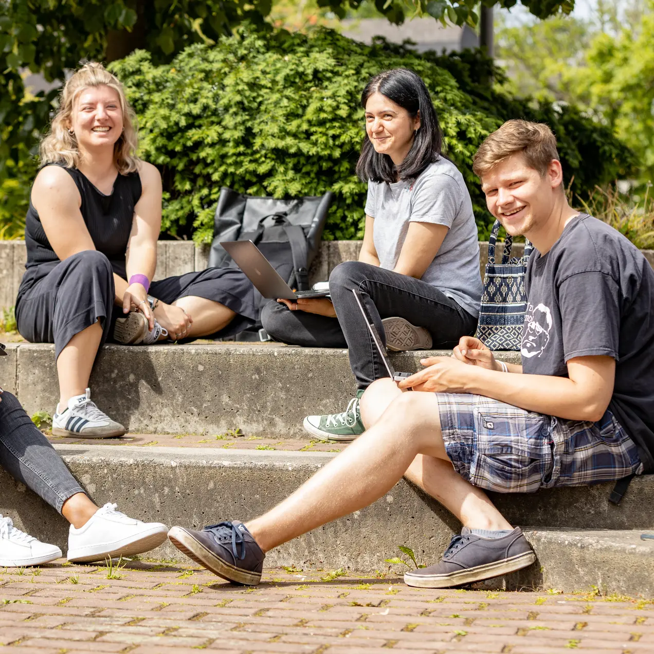Vier junge Erwachsene draußen auf einer Treppe des Hochschulgeländes mit Laptops 