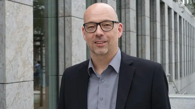 Ein Mann mit Glatze und Brille steht vor einem modernen Gebäude. Er trägt einen schwarzen Blazer und ein graues Hemd, und lächelt freundlich in die Kamera. Im Hintergrund sind große Fenster und eine steinerne Fassade sichtbar.