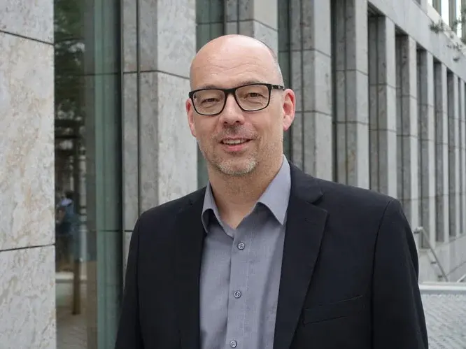 Ein Mann mit Glatze und Brille steht vor einem modernen Gebäude. Er trägt einen schwarzen Blazer und ein graues Hemd, und lächelt freundlich in die Kamera. Im Hintergrund sind große Fenster und eine steinerne Fassade sichtbar.