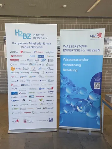 Zwei Roll-ups an einem Veranstaltungsort. Links steht "H2BZ Initiative Hessen e.V." mit dem Untertitel "Kompetente Mitglieder für ein starkes Netzwerk" und einer Liste von Logos. Rechts steht "WASSERSTOFF EXPERTISE für HESSEN" mit den Untertiteln "Wissenstransfer, Vernetzung, Beratung" und einer Grafik von Wassertropfen.