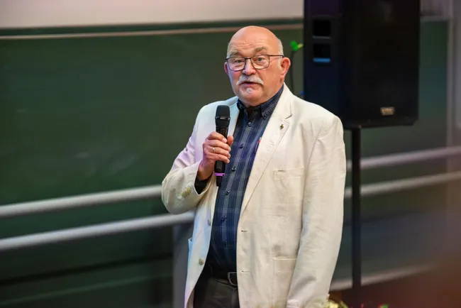 Ein älterer Mann mit Brille und einem Mikrofon steht im Vordergrund und spricht. Er trägt ein helles Jackett über einem dunklen Hemd. Im Hintergrund ist eine Tafel und ein Lautsprecher zu sehen. Die Szene vermittelt eine Atmosphäre eines Vortrags oder einer Präsentation.