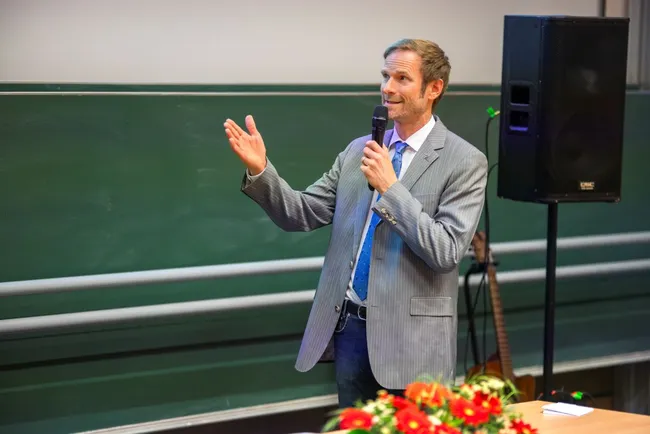 Ein Mann in einem gestreiften Anzug spricht mit einem Mikrofon in der Hand. Er gestikuliert mit seiner linken Hand, während er auf einem Podium steht, hinter dem eine begrünte Fläche sichtbar ist. Ein ­Tisch mit Blumenarrangement ist im Vordergrund zu sehen.