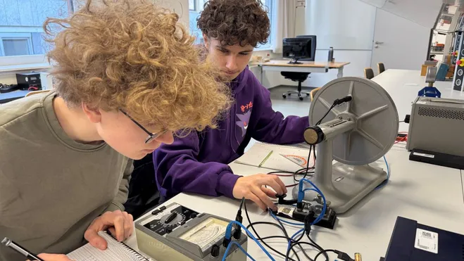 Zwei Schüler arbeiten in einem Labor. Der erste Schüler mit lockigem Haar und einer Brille macht Notizen. Der zweite Schüler am Tisch hat ebenfalls lockiges Haar und ist an einem Gerät mit einer großen Scheibe und elektrischen Komponenten beschäftigt.