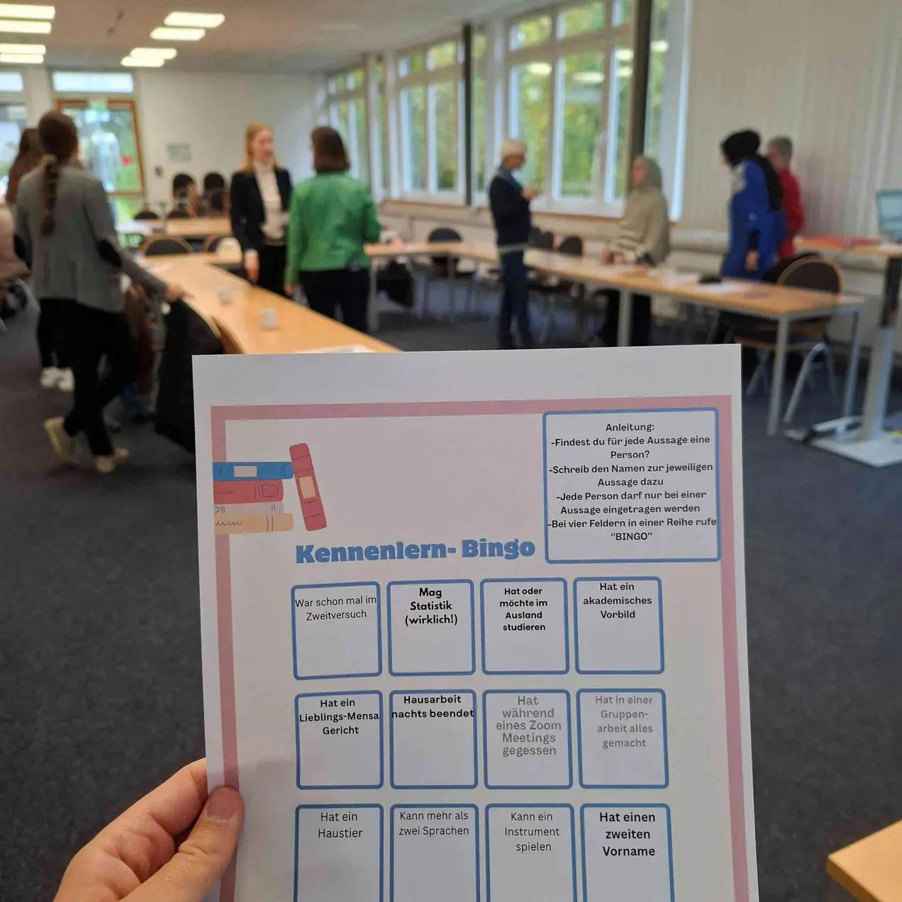 Eine Person hält ein Blatt Papier mit dem Titel „Kennenlern-Bingo“ in der Hand. Das Blatt enthält verschiedene Kästchen mit Fragen und Aussagen, die zur Interaktion und zum Kennenlernen in einer Gruppe dienen sollen. Im Hintergrund sind Teilnehmer in einem besetzten Schulungsraum zu sehen.