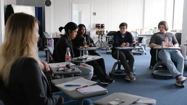 Sechs Personen sitzen in einem modernen Raum. Einige schreiben Notizen, während sie sich gegenseitig anschauen und diskutieren. Auf den Tischen stehen Getränke. Es herrscht eine konzentrierte, gemeinschaftliche Atmosphäre.