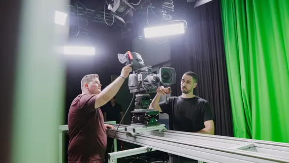 Zwei Männer arbeiten in einem Studio mit einer Videokamera auf einem Schienen-System. Einer steuert die Kamera, während der andere assistiert. Im Hintergrund ist ein grüner Vorhang sichtbar, der oft für die Produktion von Videos genutzt wird.