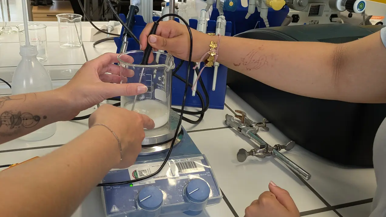 Das Bild zeigt zwei Hände, die in einem Labor an einem Experiment arbeiten. Eine Hand hält einen Kolben, während die andere eine Pipette nutzt, um Flüssigkeit hinzuzufügen. Im Hintergrund sind technische Geräte und eine Arbeitsfläche sichtbar.