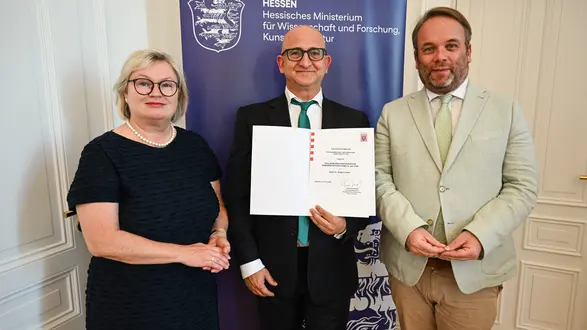 Drei Personen stehen in einem Raum vor einem Banner des Hessischen Ministeriums für Wissenschaft und Forschung. Eine Frau mit Brille und schwarzem Kleid, ein Mann, der ein Zertifikat hält, und ein Mann in heller Kleidung, der ebenfalls lächelt.