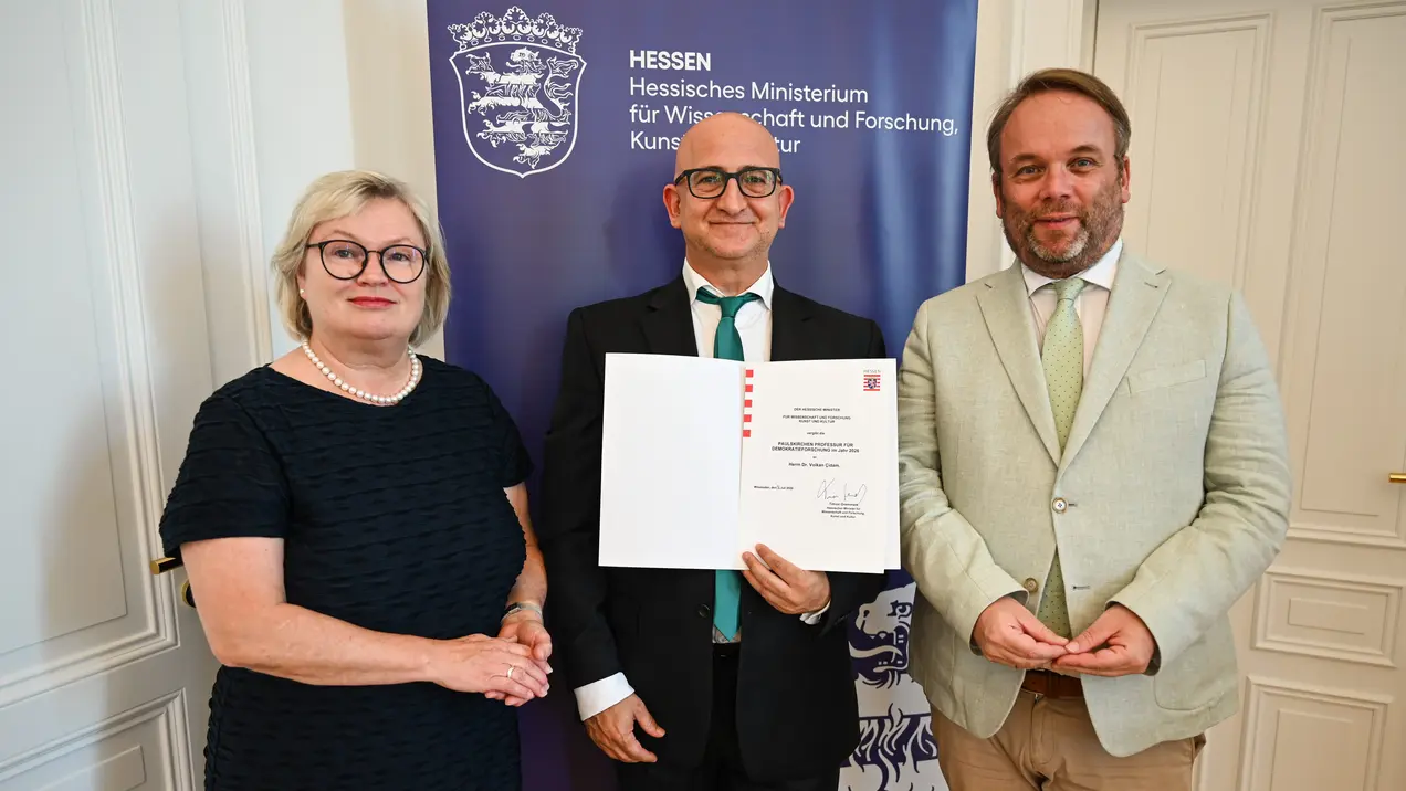 Drei Personen stehen in einem Raum vor einem Banner des Hessischen Ministeriums für Wissenschaft und Forschung. Eine Frau mit Brille und schwarzem Kleid, ein Mann, der ein Zertifikat hält, und ein Mann in heller Kleidung, der ebenfalls lächelt.