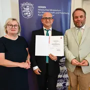 Drei Personen stehen in einem Raum vor einem Banner des Hessischen Ministeriums für Wissenschaft und Forschung. Eine Frau mit Brille und schwarzem Kleid, ein Mann, der ein Zertifikat hält, und ein Mann in heller Kleidung, der ebenfalls lächelt.
