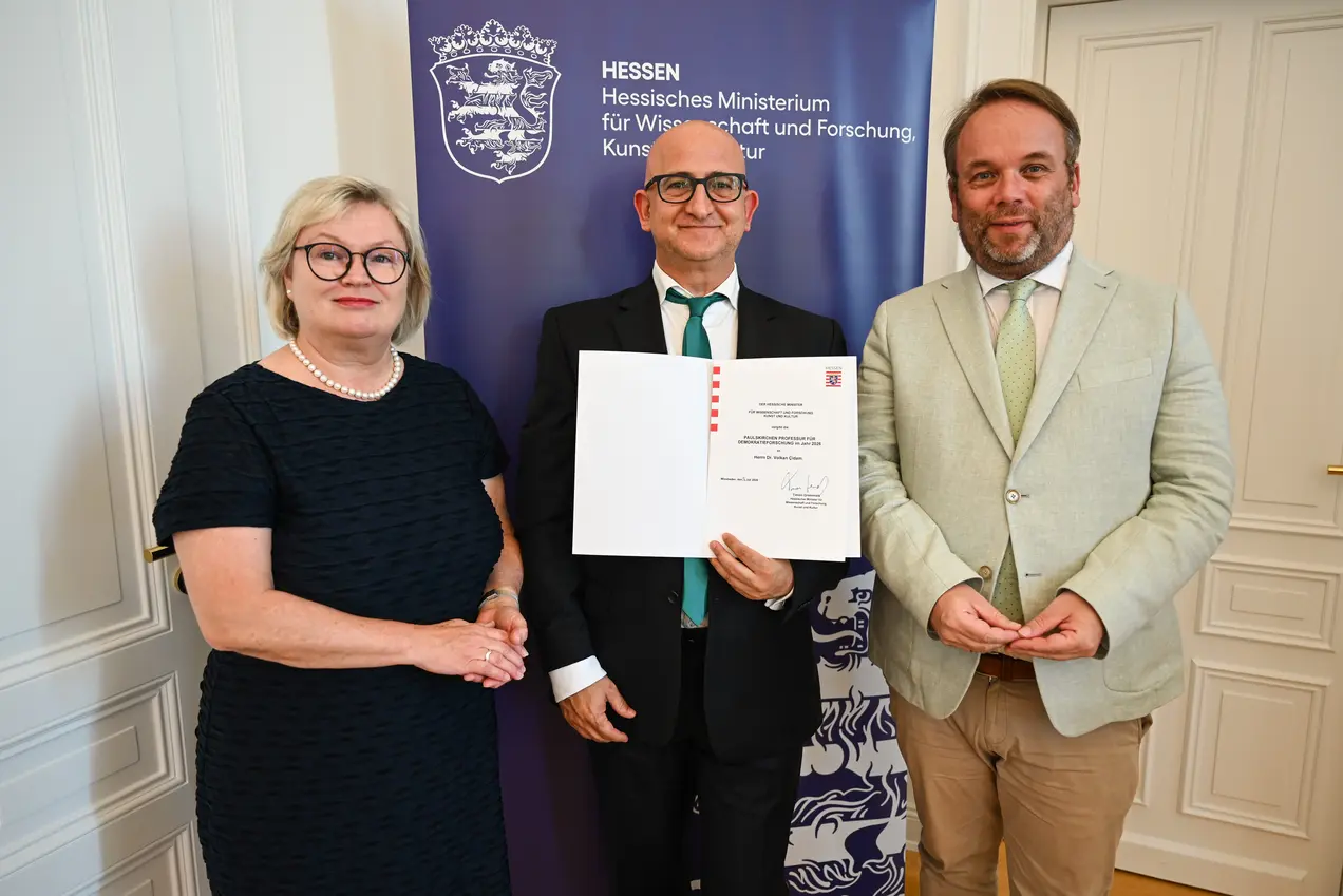Drei Personen stehen in einem Raum vor einem Banner des Hessischen Ministeriums für Wissenschaft und Forschung. Eine Frau mit Brille und schwarzem Kleid, ein Mann, der ein Zertifikat hält, und ein Mann in heller Kleidung, der ebenfalls lächelt.