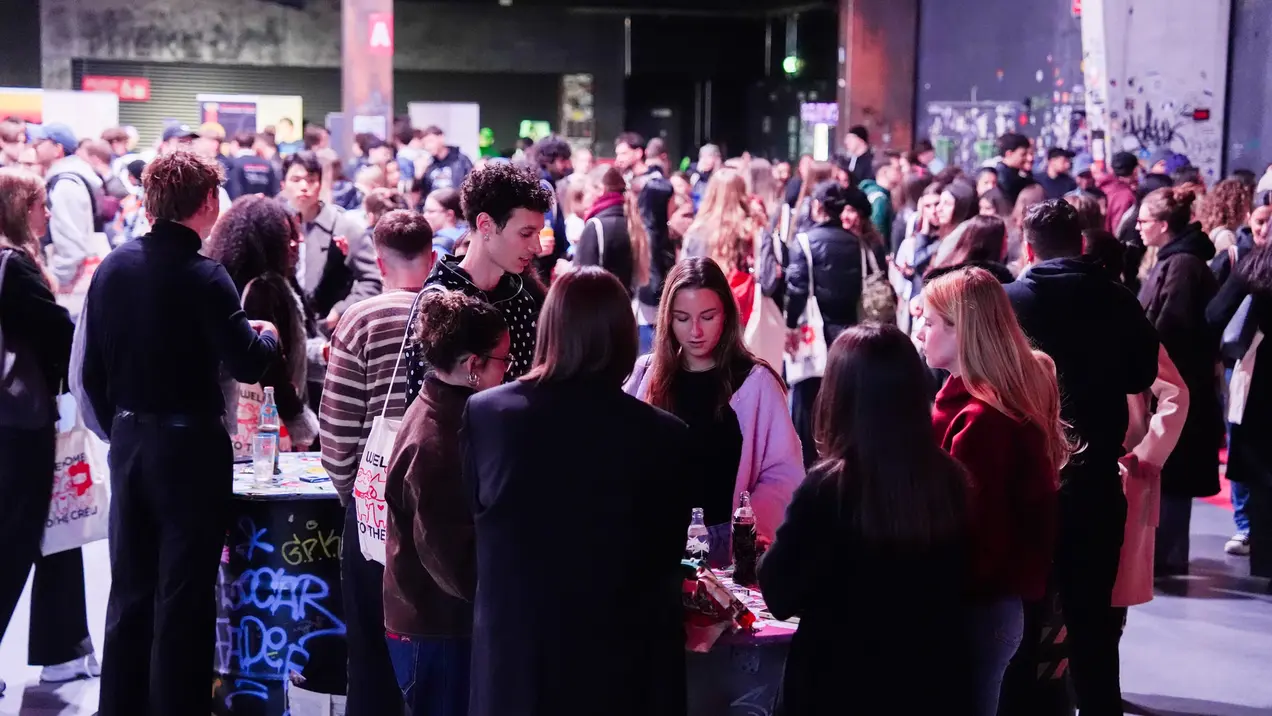 Die Aufnahme zeigt eine große Gruppe von Menschen, die sich in einem Raum versammelt haben. Einige Personen stehen im Vordergrund und interagieren miteinander, während im Hintergrund viele weitere Menschen zu sehen sind. Alle scheinen an einer Veranstaltung oder Messe teilzunehmen.