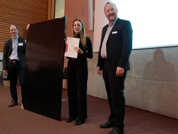 Drei Personen stehen auf einer Bühne. Denise Oberst steht in der Mitte und hält eine Auszeichnung in der Hand für die beste Leistung in allen vier Modulen der Real Estate Case Studies.