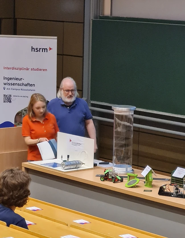 Die Abbildung zeigt zwei Personen, einen Mann mit grauen Haaren und eine Frau mit blonden Haaren, die gemeinsam an einem Tisch mit verschiedenen technischen Modellen und Präsentationsmaterialien stehen. Im Hintergrund sind Stühle und eine Schreibtafel sichtbar.