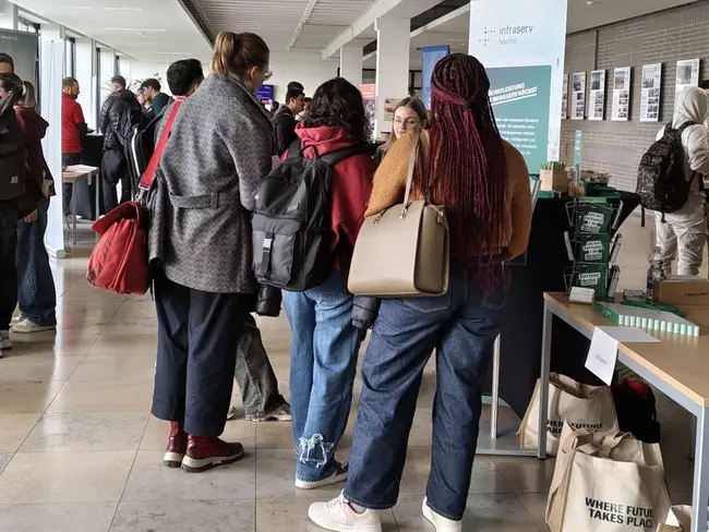 Eine Gruppe von Menschen steht in einem Flur, einige sprechen mit einer Person an einem Tisch. Im Hintergrund sind weitere Personen und Informationsstände sichtbar. Die Szene vermittelt eine Atmosphäre der Interaktion und des Austauschs.