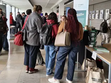 Eine Gruppe von Menschen steht in einem Flur, einige sprechen mit einer Person an einem Tisch. Im Hintergrund sind weitere Personen und Informationsstände sichtbar. Die Szene vermittelt eine Atmosphäre der Interaktion und des Austauschs.
