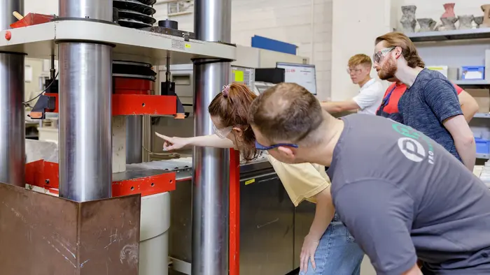 Eine Gruppe von vier Personen steht um eine hydraulische Presse im Labor für Massivbau. Eine Person zeigt auf etwas in der Presse, während die anderen aufmerksam schauen. Alle tragen Schutzbrillen, und der Raum ist gut beleuchtet, mit Werkzeugen und Materialien im Hintergrund.