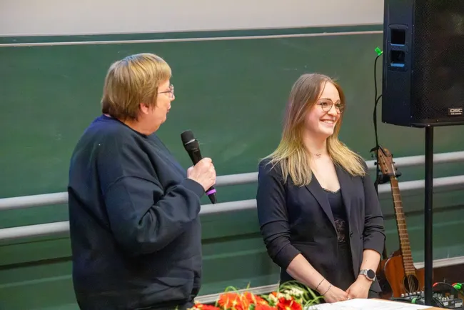Eine Person mit Brille steht lächelnd neben einer anderen Person, die ein Mikrofon hält. Beide stehen an einem Tisch, der mit Blumen dekoriert ist. Im Hintergrund ist ein Gitarrenstand zu sehen.