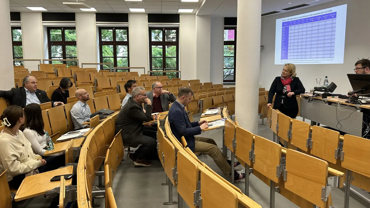 Eine Dozentin steht vorne im Hörsaal und präsentiert Informationen auf einer größeren Leinwand. Die Studenten und Zuhörer sitzen in Reihen auf Holzbänken und sind auf die Präsentation konzentriert. Der Raum ist hell und modern gestaltet.