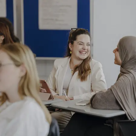Man sieht eine Gruppe von Menschen, die in einem Unterrichtsraum sitzen und zuhören. Im Vordergrund sieht man zwei Frauen, die sich anschauen und lachen.