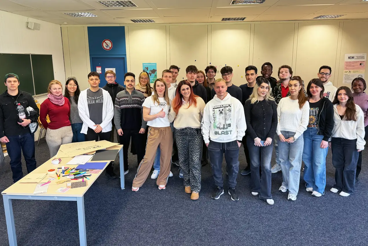 Eine Gruppe von etwa 25 Personen steht in einem Raum. Sie lächeln und stehen in mehreren Reihen. Auf einem Tisch im Vordergrund sind verschiedene Bastelmaterialien und Unterlagen sichtbar. Der Raum hat helle Wände und eine Tafel im Hintergrund.