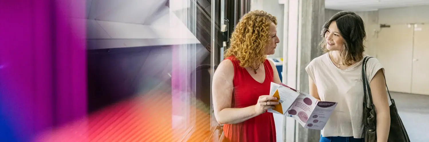 Eine Hochschulangestellte präsentiert einer Studierenden einen Flyer
