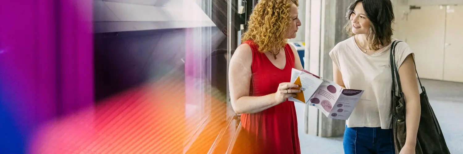 Eine Hochschulangestellte präsentiert einer Studierenden einen Flyer