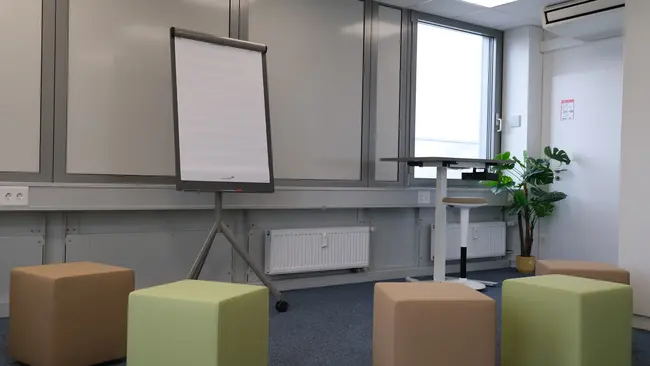 Der Raum zeigt eine besprechungsfreundliche Atmosphäre mit einem Flipchart und einem Stehpult. Rund um den Raum stehen quadratische Sitzkuben in verschiedenen Farben. Ein Fenster bringt Tageslicht herein, und eine Pflanze sorgt für eine natürliche Note.