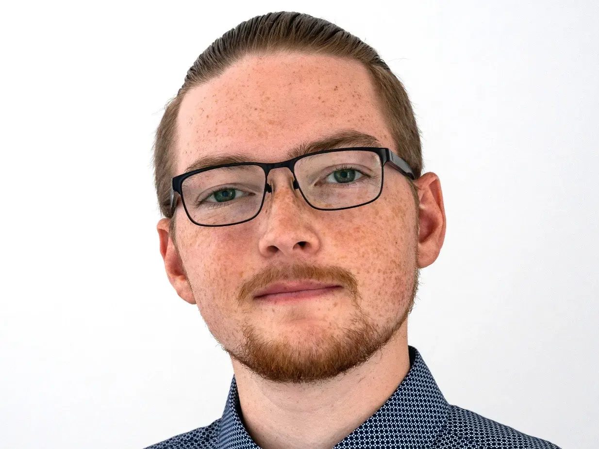 Ein junger Mann mit leichten Sommersprossen und Brille steht vor einem neutralen Hintergrund. Er trägt ein blau gemustertes Hemd und schaut direkt in die Kamera. Seine Frisur ist kurz und er hat einen gepflegten Bart.