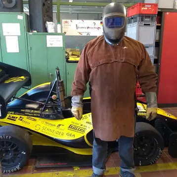 Ein Mann steht vor einem Rennwagen in einer Werkstatt. Er trägt einen Schutzanzug, Handschuhe und einen Helm. Der Wagen ist schwarz mit gelben und roten Aufklebern. Im Hintergrund sind Regale mit Werkzeugen und Ausrüstung sichtbar.