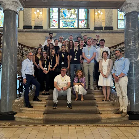 Gruppenfoto von etwa 30 Personen, die auf einer Treppe in einem historischen Gebäude stehen. Einige Personen sitzen, während andere stehen. Die Mehrzahl trägt länderspezifische Namensschilder. Im Hintergrund sind hohe Fenster mit buntem Glas zu sehen.