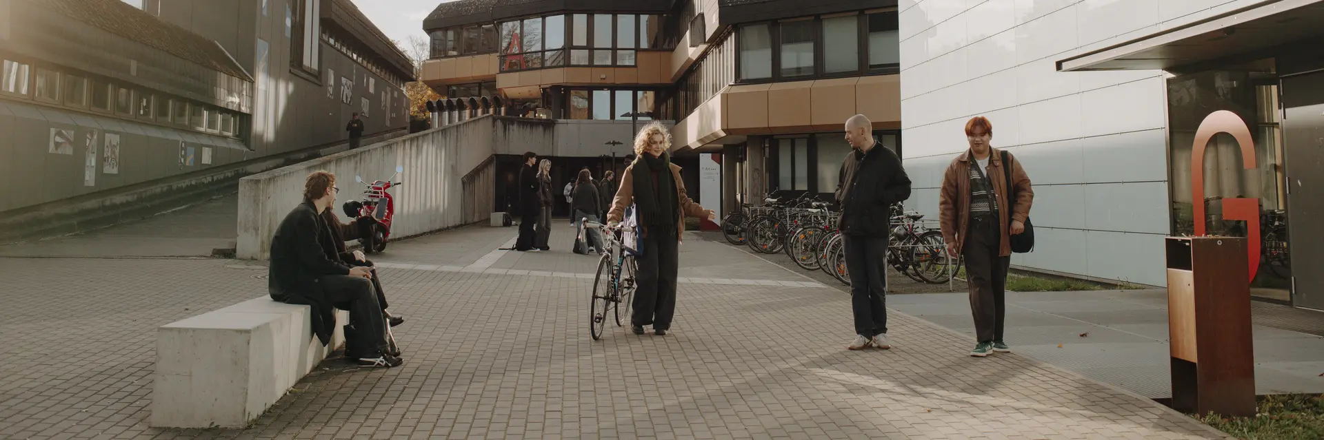 Eine Gruppe von Personen steht auf einem Platz vor modernen Gebäuden, einige stehen, andere sitzen auf einer Bank. Eine Person hält ein Fahrrad. Im Hintergrund sind weitere Fahrräder abgestellt. Der Himmel ist teilweise bewölkt und es ist tagsüber.