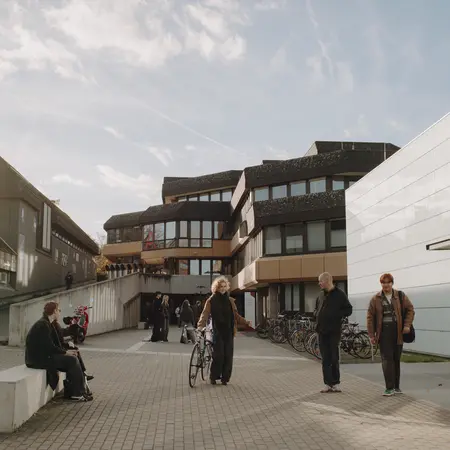 Eine Gruppe von Personen steht auf einem Platz vor modernen Gebäuden, einige stehen, andere sitzen auf einer Bank. Eine Person hält ein Fahrrad. Im Hintergrund sind weitere Fahrräder abgestellt. Der Himmel ist teilweise bewölkt und es ist tagsüber.
