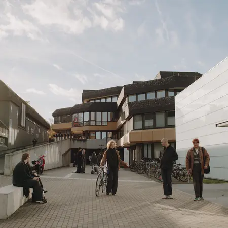 Eine Gruppe von Personen steht auf einem Platz vor modernen Gebäuden, einige stehen, andere sitzen auf einer Bank. Eine Person hält ein Fahrrad. Im Hintergrund sind weitere Fahrräder abgestellt. Der Himmel ist teilweise bewölkt und es ist tagsüber.