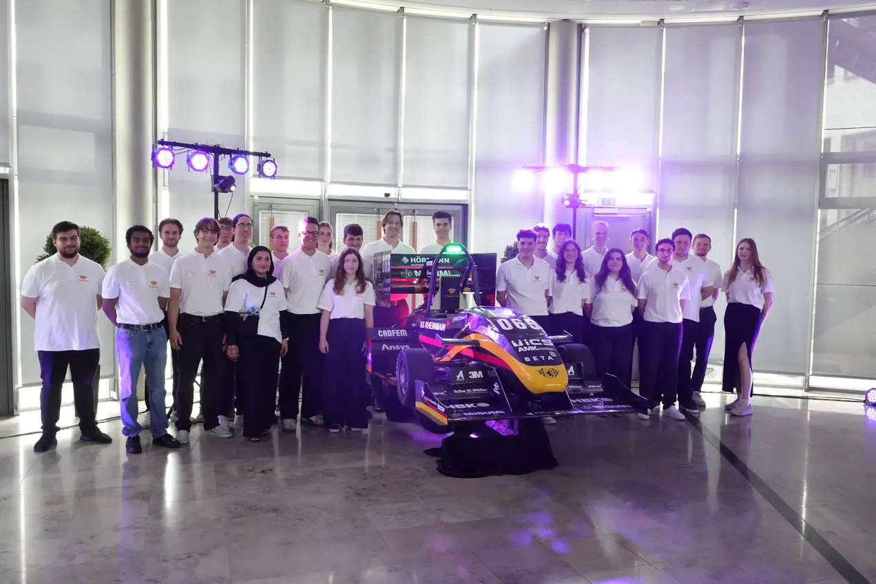 Das Team von Scuderia Mensa steht in weißen T-Shirts um ein Rennfahrzeug herum. Das Auto steht in einem modernen Innenraum, beleuchtet von verschiedenen Lichtern. 