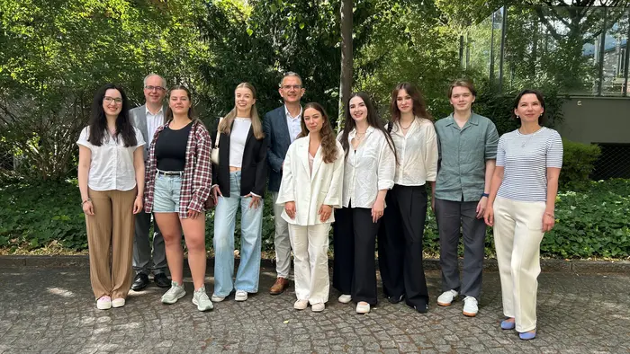 Eine Gruppe von zehn Personen steht in einem angelegten Garten. Sie posieren lächelnd in formeller und moderner Kleidung. Hinten stehen vier Personen, vorne sechs. Bäume und eine Glasarchitektur sind im Hintergrund sichtbar.