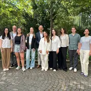 Eine Gruppe von zehn Personen steht in einem angelegten Garten. Sie posieren lächelnd in formeller und moderner Kleidung. Hinten stehen vier Personen, vorne sechs. Bäume und eine Glasarchitektur sind im Hintergrund sichtbar.