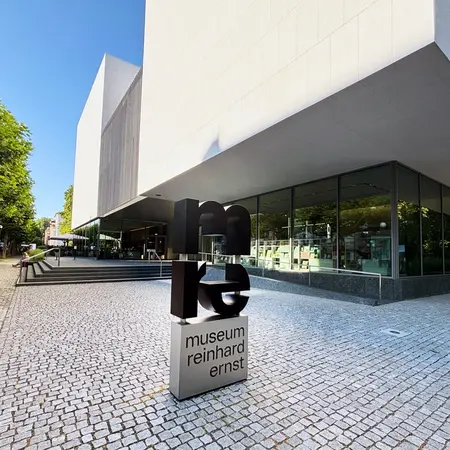 Das Bild zeigt das Gebäude des Museums Reinhard Ernst. Im Vordergrund steht ein modernes, auffälliges Schild mit dem Schriftzug „museum reinhard ernst“. Im Hintergrund sind Bäume und eine klare blaue Himmel sichtbar.