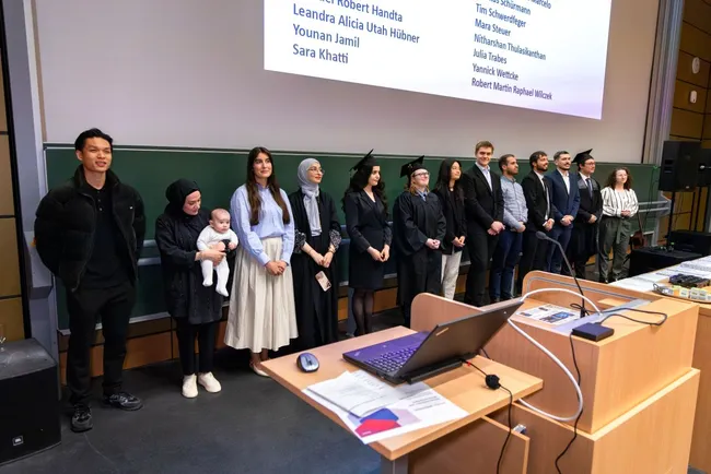 Eine Gruppe von Absolventen steht auf einer Bühne während einer Zeremonie. Einige tragen Graduation-Gowns und Hüte. Am Tisch vorne steht ein Laptop. Im Hintergrund sind Namen auf einer Leinwand zu sehen, die den Absolventen zugeschrieben sind.