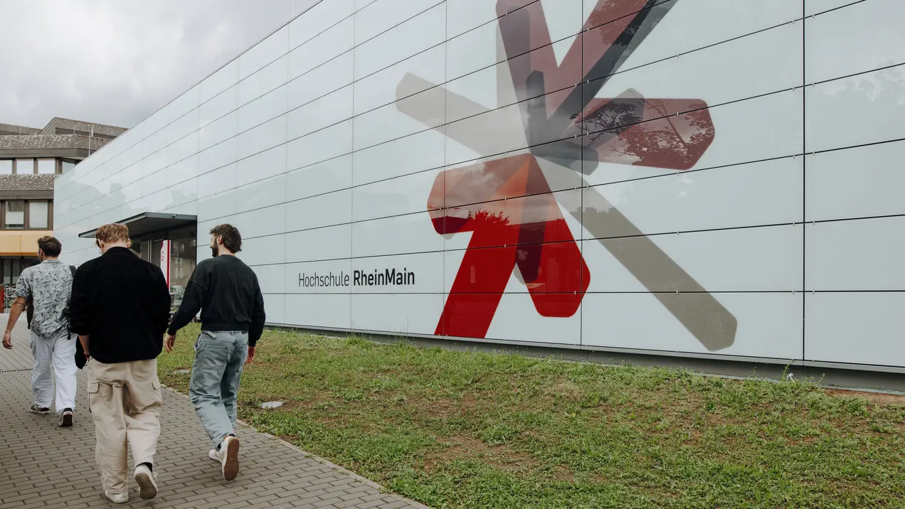 Man sieht ein modernes Gebädue mit hellblauer Fassade und der Aufschrift "Hochschule RheinMain" und einen bunten Stern aus rot, grau, schwarz und lilanen Pfeilen, die zusammenführen. Das ist das Logo der Hochschule RheinMain. Neben dem Gebäude laufen mehrere Studierende.