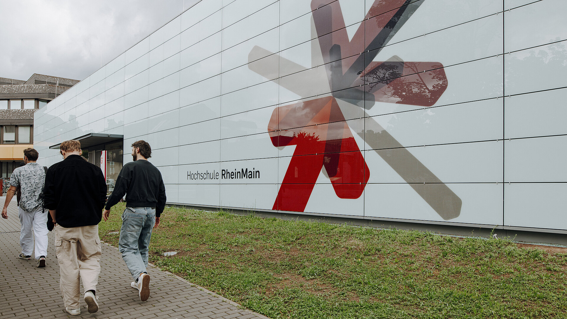 Man sieht ein modernes Gebädue mit hellblauer Fassade und der Aufschrift "Hochschule RheinMain" und einen bunten Stern aus rot, grau, schwarz und lilanen Pfeilen, die zusammenführen. Das ist das Logo der Hochschule RheinMain. Neben dem Gebäude laufen mehrere Studierende.