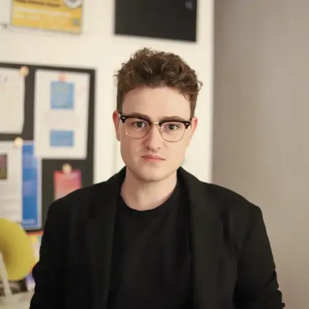 Ein junger Mann mit einer Brille und lockigem, braunem Haar sitzt in einem Raum. Er trägt einen schwarzen Blazer und blickt direkt in die Kamera. Der Hintergrund enthält einige Dokumente und Poster, die jedoch nicht im Fokus stehen.