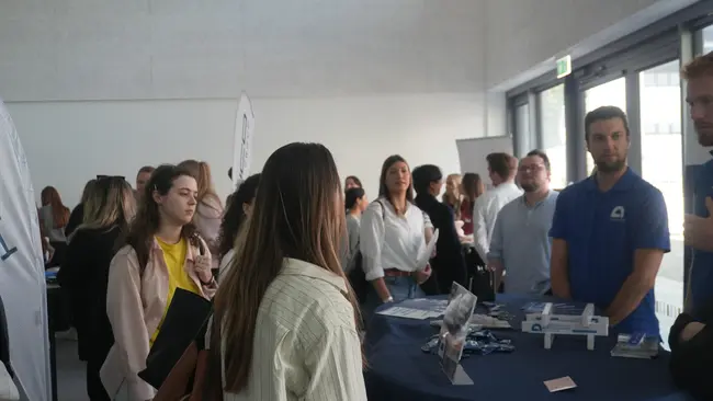 Eine Gruppe von Menschen steht an einem Stehtisch in einem Raum mit großen Fenstern. Einige Personen sprechen miteinander, während andere Informationen durchschauen. Ein Mann in einem blauen T-Shirt steht am Tisch und bietet Unterstützung an.