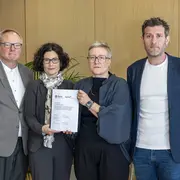 Vier Personen stehen nebeneinander in einem Raum. Eine Frau in der Mitte hält ein Dokument in der Hand. Alle schauen in die Kamera. Die Szene scheint eine wichtige Zeremonie oder ein offizielles Ereignis darzustellen.