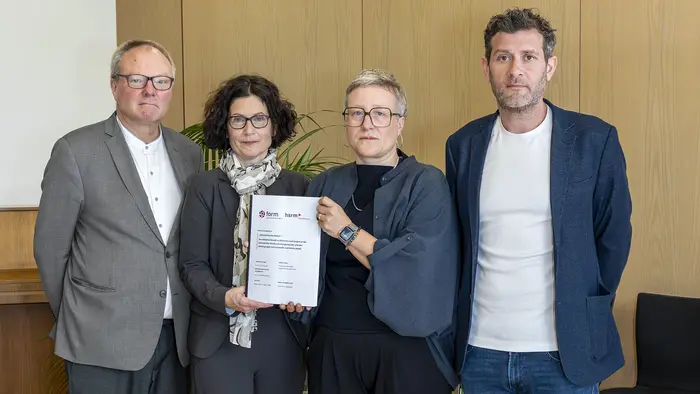 Vier Personen stehen nebeneinander in einem Raum. Eine Frau in der Mitte hält ein Dokument in der Hand. Alle schauen in die Kamera. Die Szene scheint eine wichtige Zeremonie oder ein offizielles Ereignis darzustellen.