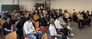 Eine Gruppe von Menschen sitzt in einem Raum, teilweise in Stuhlreihen angeordnet. Sie beobachten einen Vortrag oder eine Präsentation. Einige Zuhörer tragen Namensschilder. Die Atmosphäre wirkt konzentriert und interessiert.
