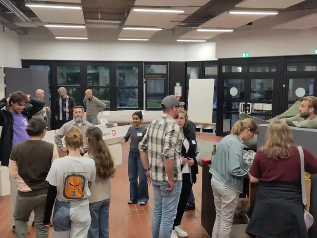 Eine Gruppe von Menschen steht in einem Raum und unterhält sich. Einige sind um Tische versammelt, während andere sich im Hintergrund bewegen. Der Raum wirkt einladend und modern, mit großen Fenstern und hellen Farben.