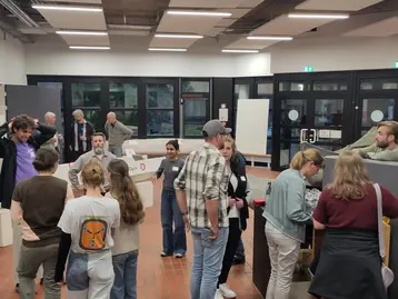 Eine Gruppe von Menschen steht in einem Raum und unterhält sich. Einige sind um Tische versammelt, während andere sich im Hintergrund bewegen. Der Raum wirkt einladend und modern, mit großen Fenstern und hellen Farben.