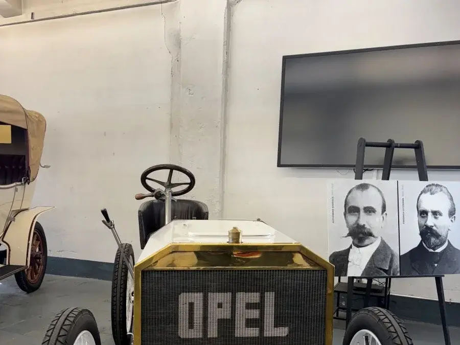 Ein historisches Auto mit der Aufschrift "OPEL" auf der Kühlerhaube steht in einem Raum. Im Hintergrund sind zwei Porträts von Männern zu sehen, die vermutlich mit der Automarke verbunden sind. Das Auto hat große, weiße Räder und eine schlichte, vintage Bauweise.